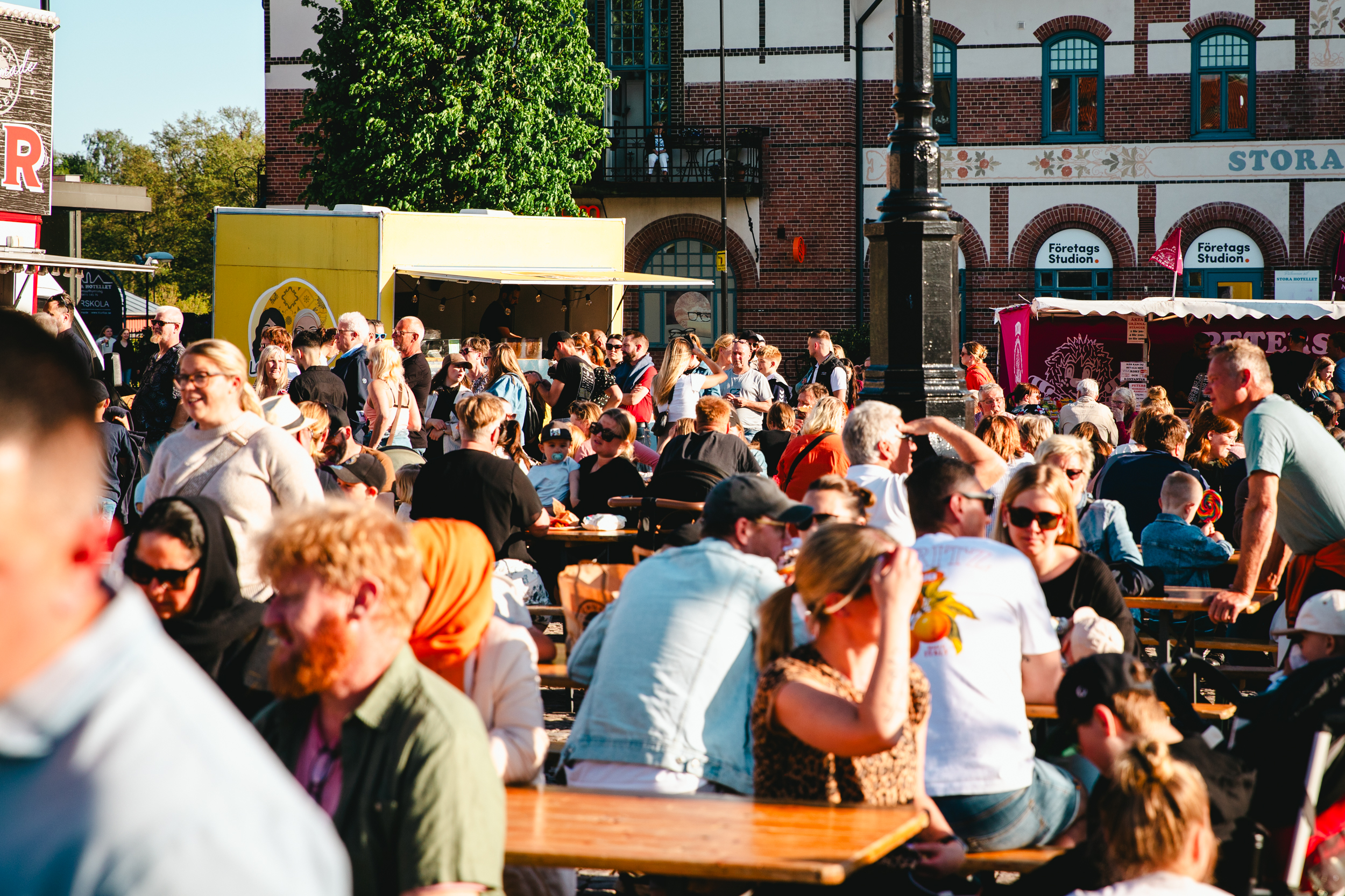 En stor folkmassa bestående av både barn och vuxna sitter utspridda vid olika bord i kvällssolen på ett torg. I bakgrunden syns också färgglada food trucks och framför dessa syns långa köer av människor.