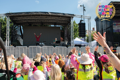 flera barn står framför en scen och sträcker upp armarna när de tittar på två personer som står på scenen och gör rörelser.