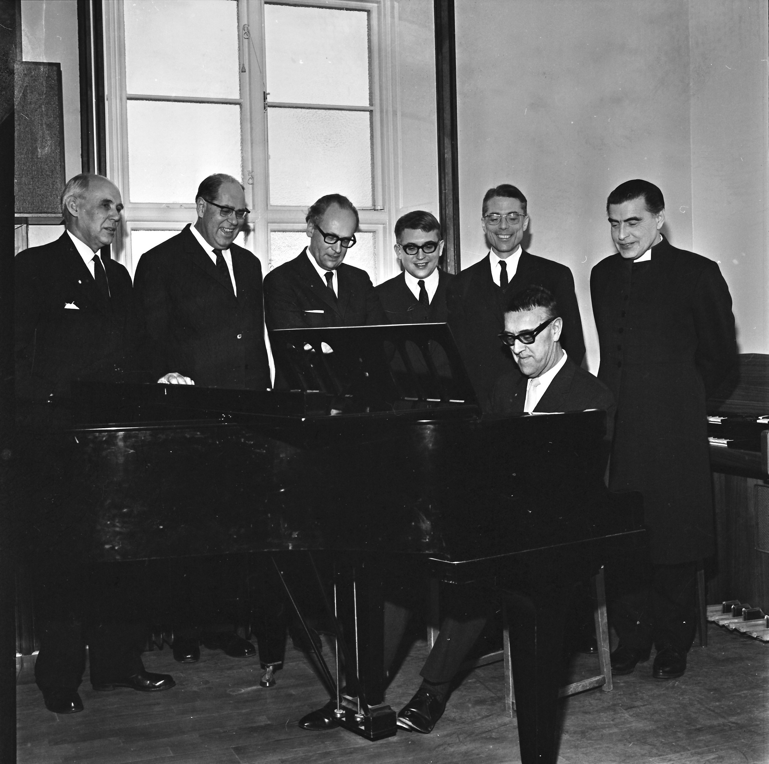 Sex kostymklädda män står upp och en sitter ner, samlade runt notställ och sjunger. Svartvit bild
