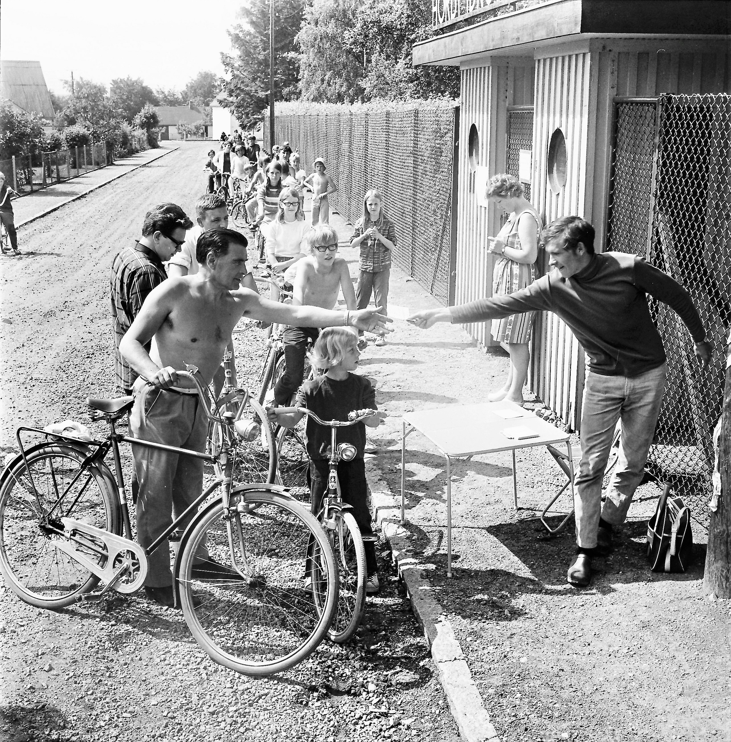 En herre sträcker på sig och delar ut något i handen till ungdomar på cykel. Utomhus, svartvit bild.