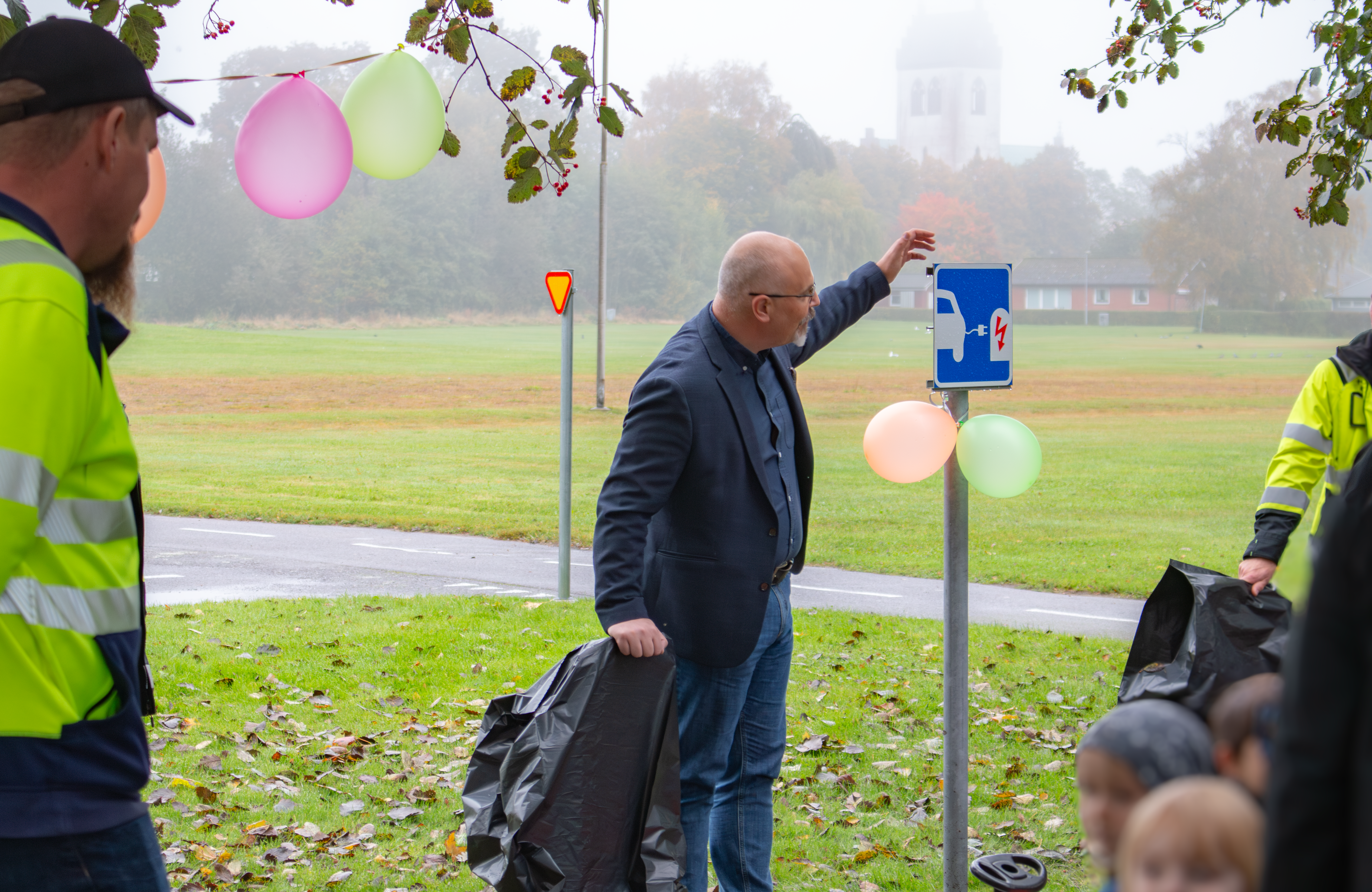 En man i kavaj står med en ballongförsedd laddstolpe i minatyr bredvid sig. I bakgrunden ser man en disig äng.