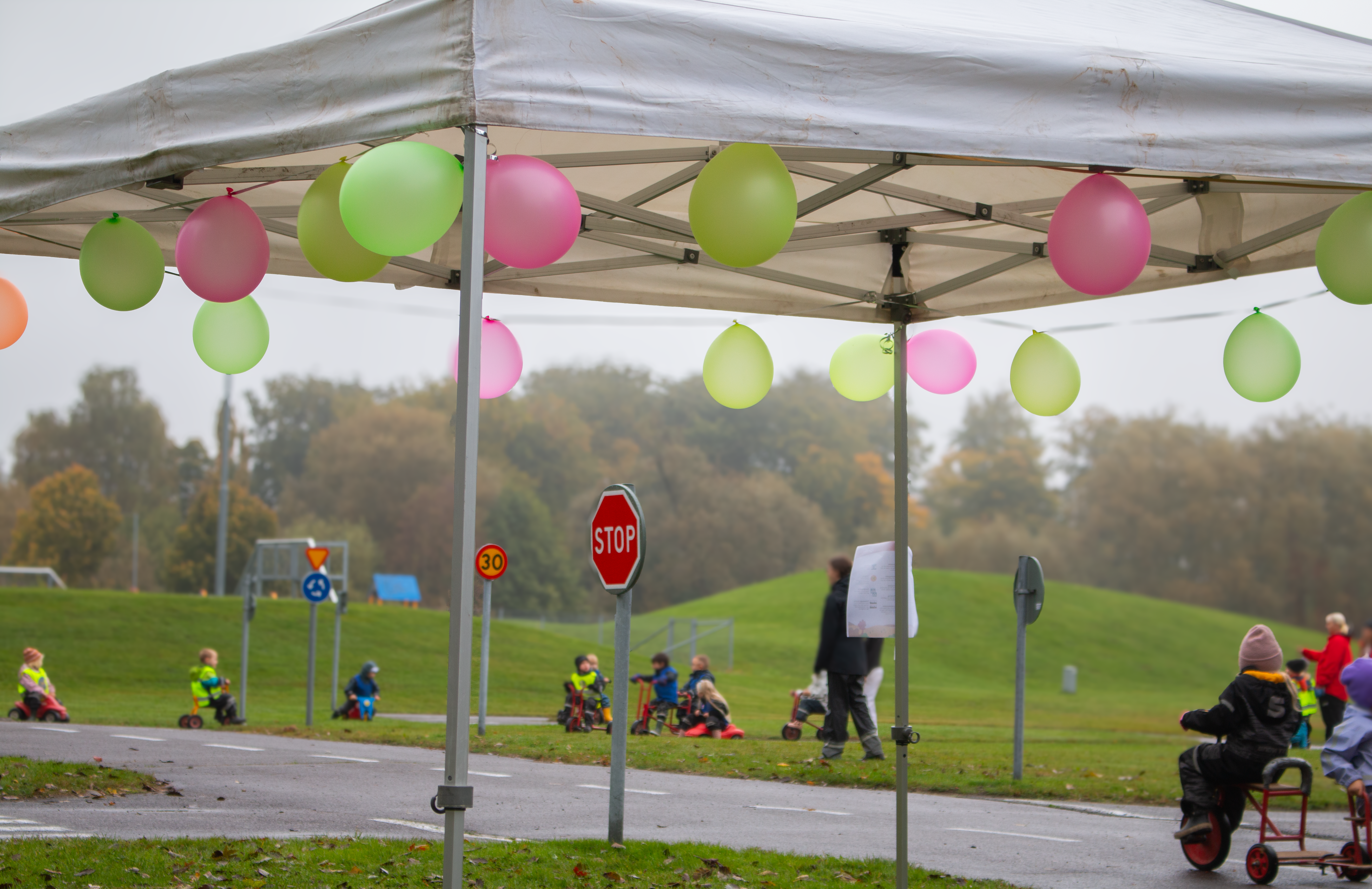 Vitt Tält prydd med färgglada ballonger i förgrunden. I Bakgrunden syns barn i gula västar cykla på trehjulingar.