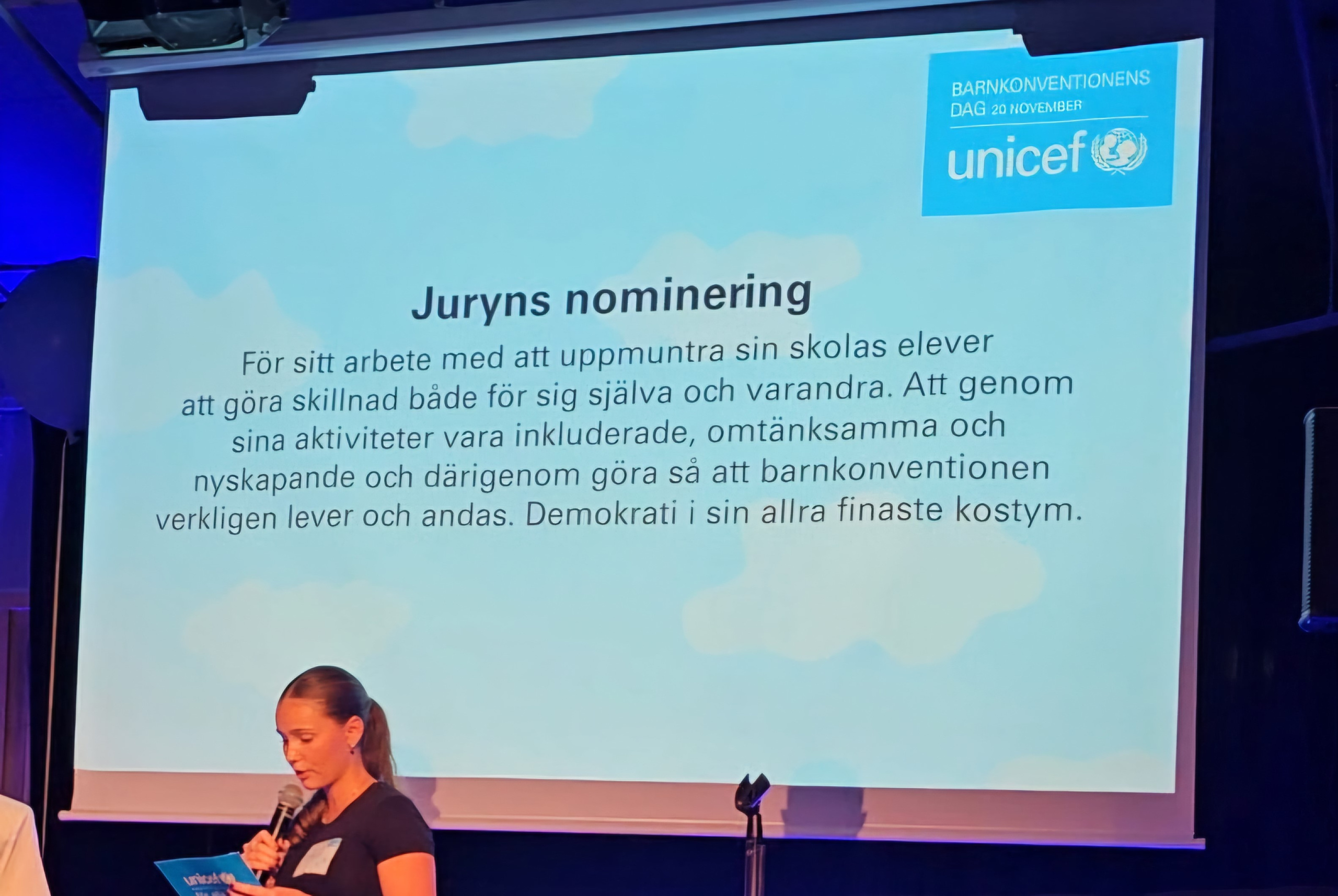 En bild på en projektorduk där juryns nominering är nedskriven. Den lyder "För sitt arbete med att uppmuntra sin skolas elever att göra skillnad både för sig själva och varandra. Att genom sina aktiviteter vara inkluderade, omtänksamma och nyskapande och därigenom göra så att barnkonventionen verkligen lever och andas. Demokrati i sin allra finaste kostym."