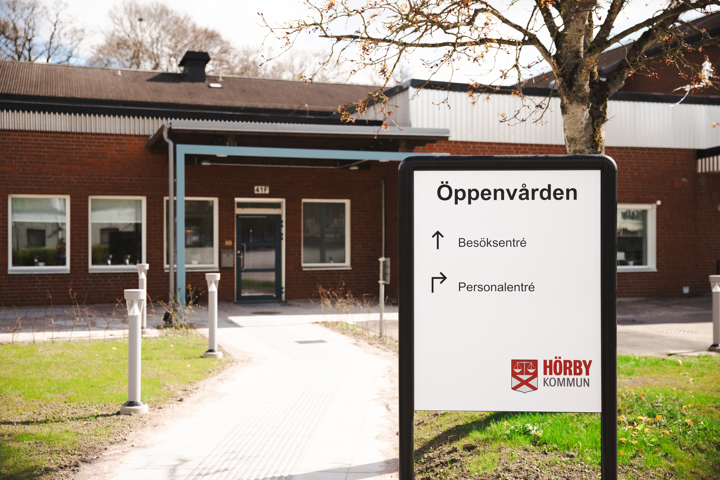 En bild tagen utomhus. I bakgrunden syns en röd tegelbyggnad och i förgrunden står en skylt med texten "Öppenvården" tillsammans med Hörby kommuns logga.